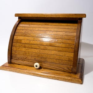 May include: A medium-brown wooden bread bin featuring a curved roll-top door with horizontal slats. The unit has a flat rectangular base and a top shelf, finished with a small, round, off-white knob centred on the front panel for opening.