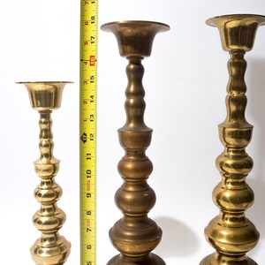 May include: Three brass candle holders of varying heights. The tallest is approximately 18 inches tall, the middle one is about 14 inches, and the shortest is around 10 inches. Each has a flared top and a decorative, bulbous stem.