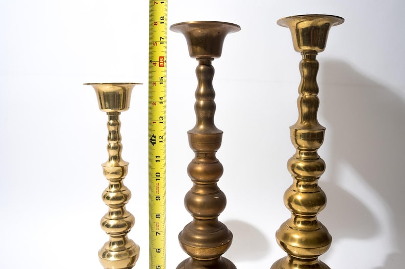 May include: Three brass candle holders of varying heights. The tallest is approximately 18 inches tall, the middle one is about 14 inches, and the shortest is around 10 inches. Each has a flared top and a decorative, bulbous stem.