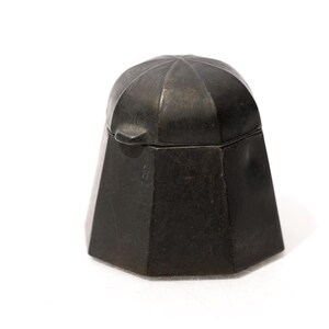 May include: A dark gray, matte-finished metal container with a faceted, hexagonal base and a matching domed lid. The lid features a small, protruding tab for lifting, and the overall structure has a solid, geometric appearance with clean lines.