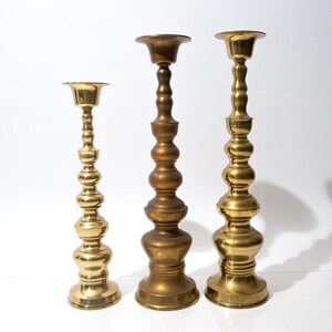 May include: Three brass candlesticks of varying heights stand against a white background. The candlesticks have ornate, bulbous designs and are finished in a shiny gold color. They are suitable for holding candles.