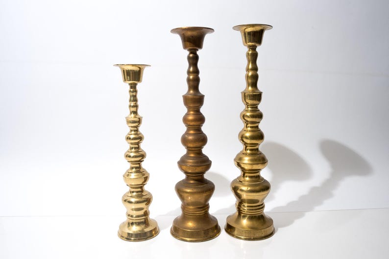 May include: Three brass candlesticks of varying heights stand against a white background. The candlesticks have ornate, bulbous designs and are finished in a shiny gold color. They are suitable for holding candles.