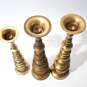 May include: Three antique brass candle holders of varying heights. Each holder has a wide, shallow cup for the candle and a decorative, tiered base. The brass has a warm, aged patina, suggesting vintage pieces. The candle holders are arranged on a white surface.