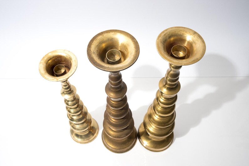 May include: Three antique brass candle holders of varying heights. Each holder has a wide, shallow cup for the candle and a decorative, tiered base. The brass has a warm, aged patina, suggesting vintage pieces. The candle holders are arranged on a white surface.