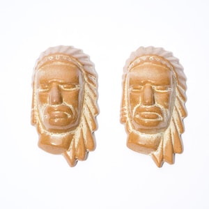May include: Two ceramic sculptures of Native American heads, featuring detailed facial expressions and headdresses. The sculptures are a warm brown colour with lighter accents, set against a white background.