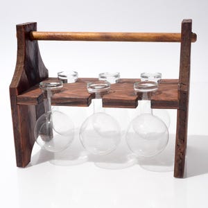 May include: A dark wooden wine glass rack holding six clear wine glasses. The rack has a handle and is designed to hold the glasses upside down. The wood is a rich, dark brown colour.