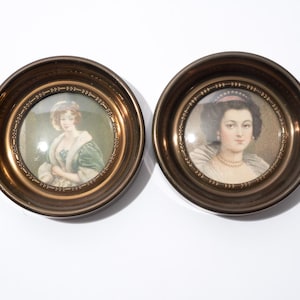 May include: Two antique portrait miniatures in round, ornate, gold-toned frames. The portraits depict women in period clothing, one in a green and white dress, the other in a white dress with a pearl necklace. The frames have a decorative edge.
