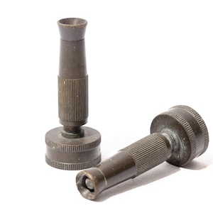 Puede incluir: Dos boquillas de manguera antiguas de color bronce. Una está de pie, la otra está tumbada de lado. Ambas tienen un cuerpo cilíndrico con detalles acanalados y una parte superior ensanchada. Sobre fondo blanco.