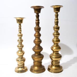 May include: Three brass candlesticks of varying heights stand against a white backdrop. The candlesticks have a classic design with a round base, a long, decorative stem, and a cup-shaped top to hold a candle.