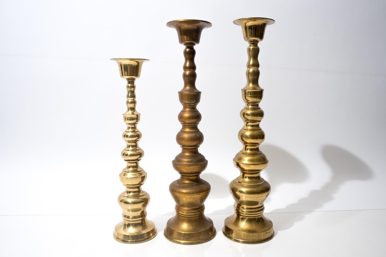 May include: Three brass candlesticks of varying heights stand against a white backdrop. The candlesticks have a classic design with a round base, a long, decorative stem, and a cup-shaped top to hold a candle.