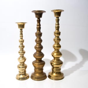 May include: Three brass candlesticks of varying heights stand against a white background. The candlesticks have a classic, ornate design with a gold finish. The tallest candlestick is approximately 12 inches tall.
