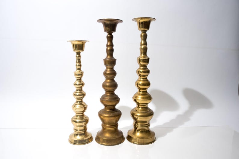 May include: Three brass candlesticks of varying heights stand against a white background. The candlesticks have a classic, ornate design with a gold finish. The tallest candlestick is approximately 12 inches tall.