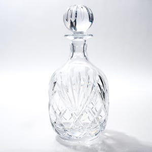 May include: A clear glass decanter with a decorative stopper. The decanter features a cut glass design and a rounded base. The stopper is a round shape with vertical lines. The design is elegant and classic.