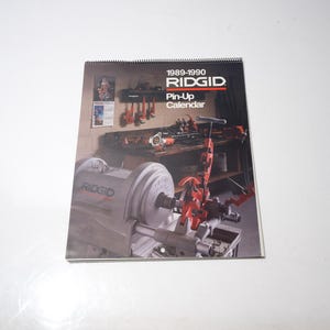 Peut inclure: Un calendrier Pin-Up RIDGID 1989-1990. La couverture pr&eacute;sente une sc&egrave;ne d'atelier avec des outils et des machines. Le calendrier est reli&eacute; en spirale et porte le logo RIDGID. Le fond est blanc.