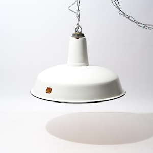 Könnte beinhalten: Eine weiße, emaillierte Metall-Hängeleuchte mit einem breiten, kuppelförmigen Schirm und einem schwarzen Rand. Die Lampe ist an einer silberfarbenen Metallkette aufgehängt und hat ein kleines, rechteckiges braunes Etikett am Schirm.