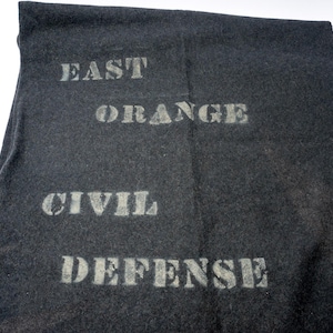 Peut inclure: Textile gris foncé avec les mots "EAST ORANGE CIVIL DEFENSE" pochoirés en gris clair. Le tissu semble être une matière lourde, peut-être de la laine ou du feutre, avec une surface légèrement texturée.