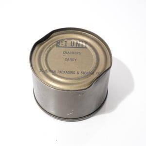 Puede incluir: Una lata de metal de color verde oliva con la parte superior ligeramente abollada. La etiqueta dice "B-1 UNIT" encima de "CRACKERS" y "CANDY". La lata es de "SOUTHERN PACKAGING & STORAGE CO."
