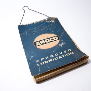 Puede incluir: Guía de lubricación Amoco Approved de 1957. La cubierta azul presenta texto blanco y el logotipo de Amoco en un óvalo beige. Encuadernación en espiral con colgador metálico. Páginas envejecidas.