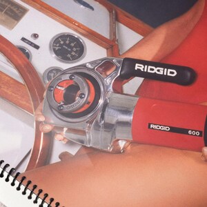 Peut inclure: Un outil de filetage de tuyaux RIDGID 600 rouge et argent&eacute;. L'outil a une poign&eacute;e noire avec le logo RIDGID. L'outil est tenu pr&egrave;s d'un volant en bois et d'un tableau de bord blanc avec des jauges.