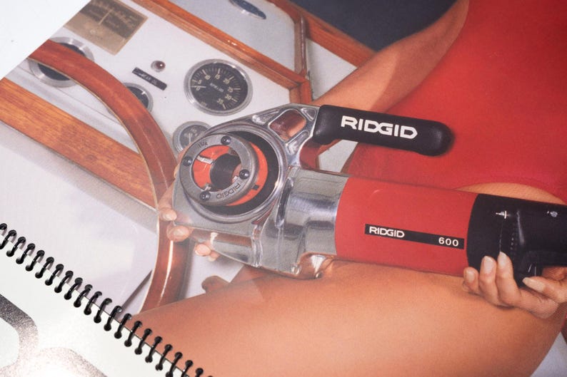 Peut inclure: Un outil de filetage de tuyaux RIDGID 600 rouge et argent&eacute;. L'outil a une poign&eacute;e noire avec le logo RIDGID. L'outil est tenu pr&egrave;s d'un volant en bois et d'un tableau de bord blanc avec des jauges.