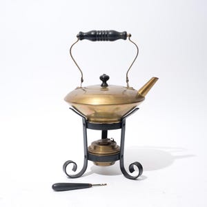 May include: Antique brass tea kettle with a black handle and stand. The kettle has a rounded body, a spout, and a lid. The stand is black with decorative scrollwork. A small black-handled tool rests in front.