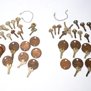 May include: A collection of vintage keys and round metal tags with text. The keys are various sizes and shapes, with a mix of silver and bronze tones. The tags are a dark bronze color, some with attached keys.