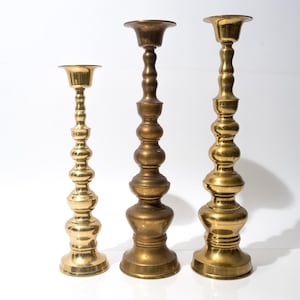 May include: Three brass candlesticks of varying heights stand against a white background. The candlesticks have a classic design with a round base, a long, ornate stem, and a cup-shaped top. The tallest candlestick is approximately 12 inches tall.