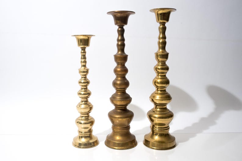 May include: Three brass candlesticks of varying heights stand against a white background. The candlesticks have a classic design with a round base, a long, ornate stem, and a cup-shaped top. The tallest candlestick is approximately 12 inches tall.