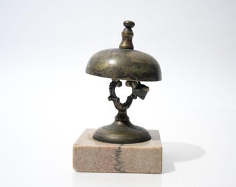 Op de afbeelding: Een antieke messing bureaubel met een gewelfde bovenkant en een sierlijke basis, gemonteerd op een vierkante marmeren basis. De bel heeft een kleine klepel en een donkere, verouderde patina, wat wijst op zijn vintage. Het is een decoratief item.