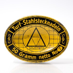 Könnte beinhalten: Ovale Dose mit schwarzem Text und gelbem Hintergrund. Der Text lautet "Turf-Stahlstecknadeln" und "50 Gramm netto No 7". Ein schwarzes Dreieck befindet sich in einem gelben Kreis. Die Dose ist wahrscheinlich für Nähnadeln.
