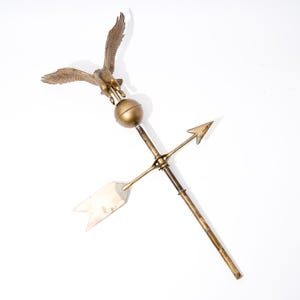 May include: A vintage brass weathervane featuring a detailed eagle with outstretched wings atop a sphere, with directional arrows. The weathervane is made of brass and has a weathered appearance.