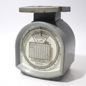 Puede incluir: Balanza postal Pelouze "A" Line, modelo A-10-12, de color gris. La esfera circular muestra el peso y las tarifas postales. La plataforma superior es de metal.