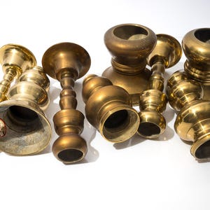 May include: A collection of antique brass candle holders in various shapes and sizes. The candle holders have a warm, golden hue and are arranged on a white surface. Some have a bell-shaped base, while others have a more rounded design.