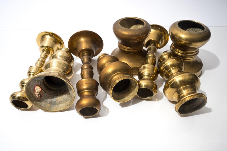 May include: A collection of antique brass candle holders in various shapes and sizes. The candle holders have a warm, golden hue and are arranged on a white surface. Some have a bell-shaped base, while others have a more rounded design.