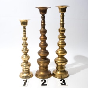 May include: Three brass candle holders of varying heights. The candle holders have a decorative, bulbous design. The tallest candle holder is approximately 12 inches tall. The other two are shorter, with the smallest being about 8 inches tall. Each has a circular base and a cup-shaped top.