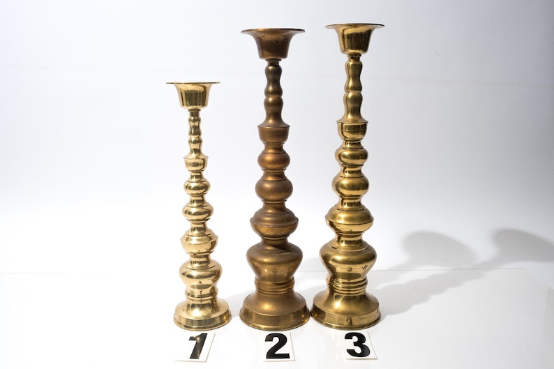 May include: Three brass candle holders of varying heights. The candle holders have a decorative, bulbous design. The tallest candle holder is approximately 12 inches tall. The other two are shorter, with the smallest being about 8 inches tall. Each has a circular base and a cup-shaped top.