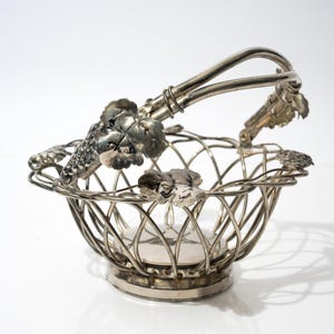 May include: Silver-toned metal basket with a handle and a lattice design. The basket is decorated with grape and leaf motifs. The handle is arched, and the basket sits on a round base. A decorative serving piece.