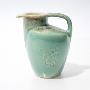 May include: A turquoise ceramic pitcher with a handle and spout. The pitcher has a leaf design on the front and a brown rim. The pitcher is approximately 7 inches tall.