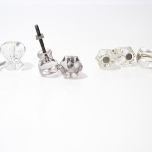 May include: A collection of clear glass cabinet knobs in various shapes. Some knobs have screws or bolts attached. The knobs are faceted and reflect light, creating a sparkling effect. The hardware is suitable for furniture restoration or DIY projects.