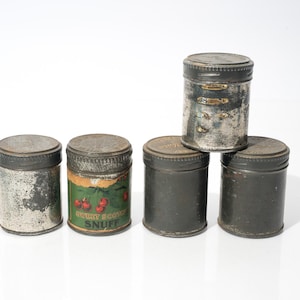 May include: A collection of five antique metal snuff tins with screw-on lids. The tins vary in color, from silver to dark gray, with signs of wear and tear. One tin features a label with cherries and the text "Sweet Scotch Snuff."