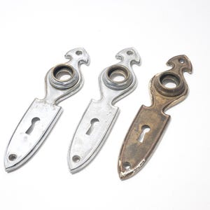 May include: Three vintage metal door escutcheons. Two are silver-toned, and one is bronze-toned. Each has a keyhole and a decorative top with a circular opening. The escutcheons are on a white background.