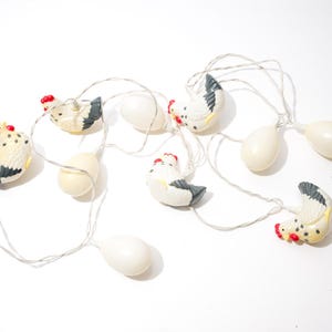 Könnte beinhalten: Dekorative Lichterkette mit kleinen Hühner- und Eierornamenten. Die Hühner sind weiß mit schwarzen und gelben Akzenten und roten Kämmen. Die Eier sind cremeweiß und an einem silbernen Draht befestigt.