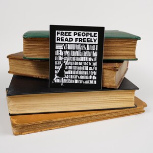 3.4" x4" matte rectangular sticker made from a linocut print, which has the text "Free people read freely" in block capitals above a person in a dress reaching to take a book from a large bookshelf. The sticker is propped on a stack of old books.