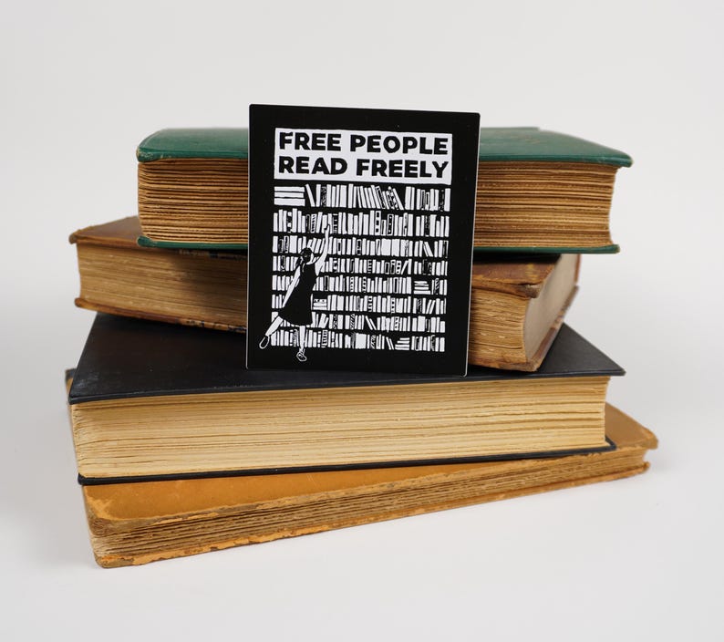 3.4" x4" matte rectangular sticker made from a linocut print, which has the text "Free people read freely" in block capitals above a person in a dress reaching to take a book from a large bookshelf. The sticker is propped on a stack of old books.
