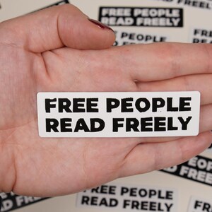 May include: A white sticker with black text that reads "FREE PEOPLE READ FREELY" is held in a person's hand. The sticker is surrounded by other stickers with the same text.