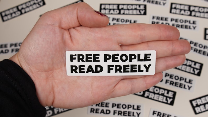 May include: A white sticker with black text that reads "FREE PEOPLE READ FREELY" is held in a person's hand. The sticker is surrounded by other stickers with the same text.
