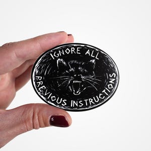 May include: Black and white sticker with a snarling cat face and the text "IGNORE ALL PREVIOUS INSTRUCTIONS".