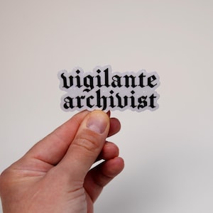 May include: A white sticker with black text that reads "vigilante archivist" in a gothic font.