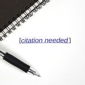 May include: A black pen with a silver clip and a clear barrel lies on a white surface next to a black spiral bound notebook. The text "[citation needed]" is written in blue ink on the white surface.