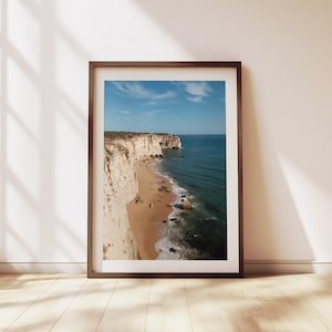 May include: A photograph of a white cliff face along a sandy beach with blue ocean waves crashing on the shore. The sky is blue with white clouds.